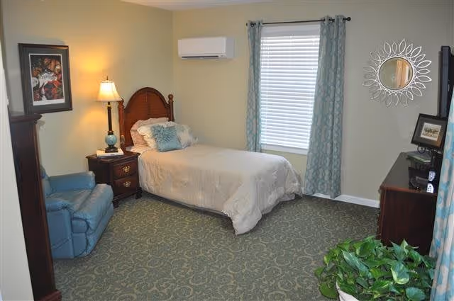 A cozy bedroom with a single bed featuring a wooden headboard, white bedding, and decorative pillows. Next to the bed is a wooden nightstand with a lamp and a framed picture on the wall above it. A blue recliner chair is positioned near the nightstand. The room has a window with blue patterned curtains, a wall-mounted air conditioning unit above the bed, a wooden dresser with a framed photo and a decorative round mirror on the wall. There is a green potted plant in the corner of the room and the floor is covered with a patterned carpet.