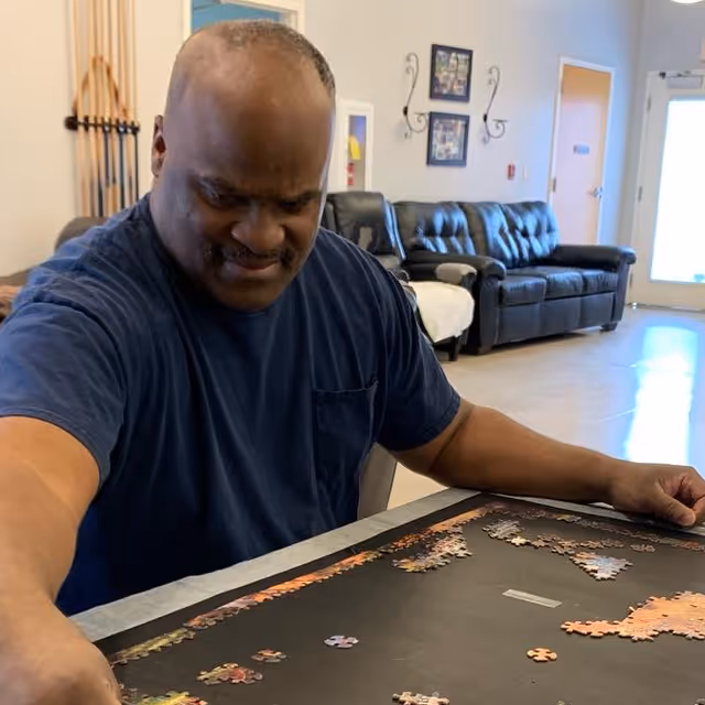 A man in a navy blue shirt is sitting at a table working on a jigsaw puzzle in a room with a black leather couch and framed pictures on the wall in the background.