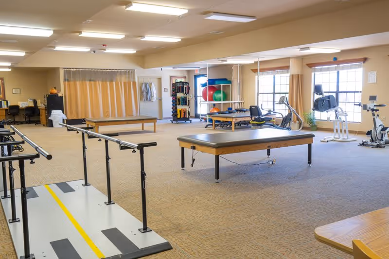 A spacious physical therapy room with parallel bars for walking exercises, two treatment tables, exercise balls, resistance bands, stationary exercise bikes, and large windows letting in natural light.