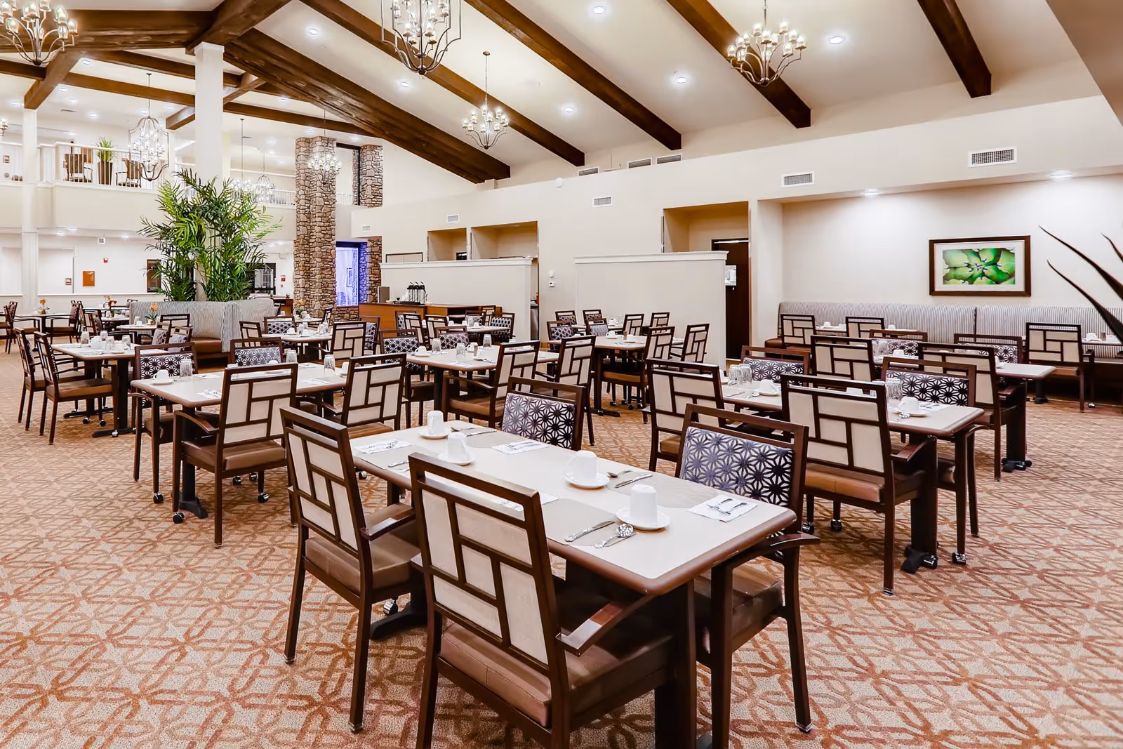 Spacious dining room with multiple tables and chairs arranged neatly. Each table is set with white cups, saucers, and silverware. The room features high ceilings with exposed wooden beams, chandeliers, and a stone pillar. There are plants and framed artwork on the walls, creating a warm and inviting atmosphere.