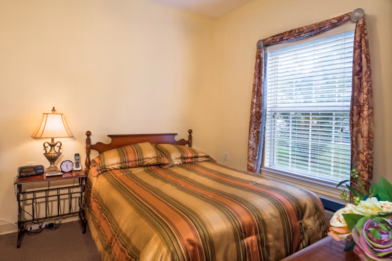 A cozy bedroom with a wooden bed frame and a striped bedspread in shades of brown, green, and gold. There is a bedside table with a lamp, clock, and phone on the left side of the bed. A window with white blinds and floral curtains is on the right wall, letting in natural light. A small bouquet of flowers is partially visible on a piece of furniture in the foreground.