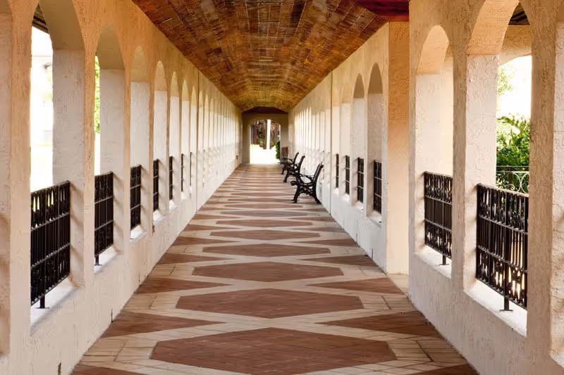 A long covered outdoor corridor with arched openings on both sides, decorative iron railings, patterned tile flooring, and several benches along the right side under a wooden ceiling.
