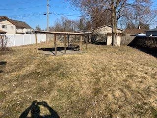 A fenced backyard with dry grass, a small wooden pergola-style pavilion in the center, neighboring houses, and a utility pole under a clear blue sky.