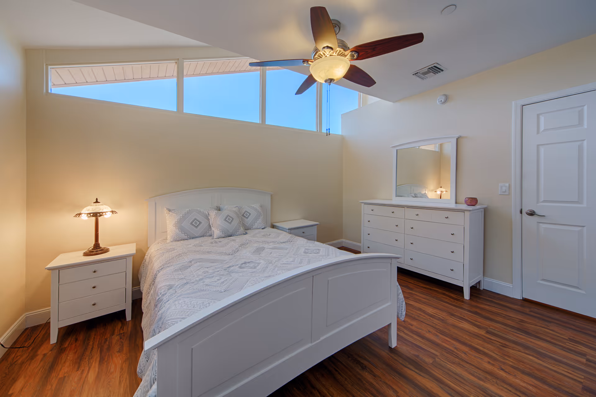 Bright bedroom with a white wooden bed, matching nightstands and dresser, ceiling fan, and high horizontal windows.