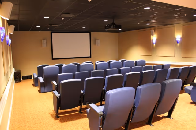 A small theater room with multiple rows of blue cushioned seats facing a blank projection screen mounted on a beige wall. The room has a carpeted floor and soft wall lighting with blue sconces.