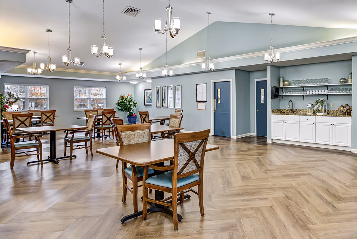 A spacious dining room with multiple wooden tables and chairs arranged neatly. The room has light blue walls, large windows letting in natural light, and several modern chandeliers hanging from the ceiling. There is a counter area with white cabinets, granite countertop, shelves with glasses and cups, and decorative items. The floor has a wood pattern design.