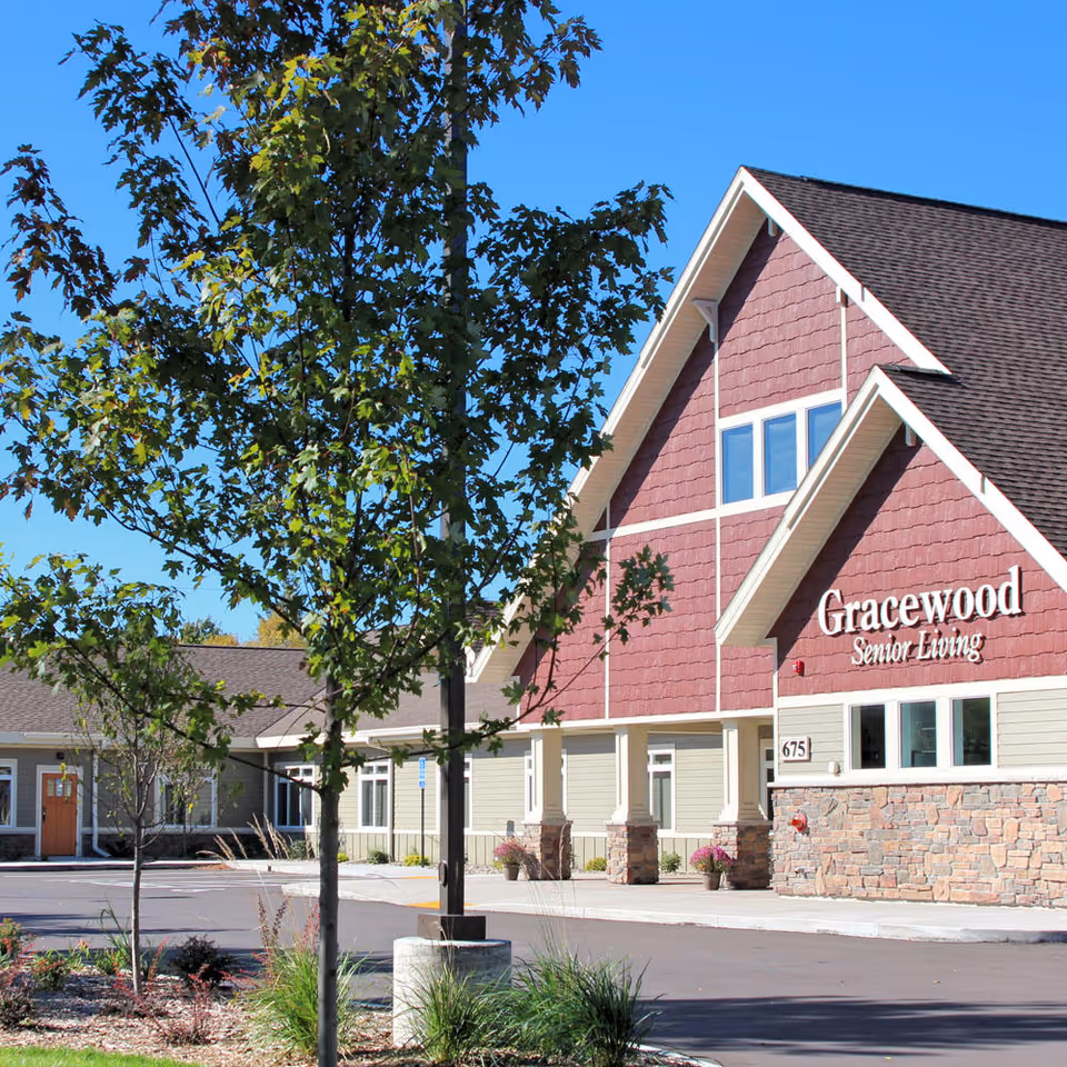 Exterior view of Gracewood Senior Living building with a red and beige facade, stone accents, and a peaked roof under a clear blue sky. There are trees and landscaping in front of the building and a paved driveway or parking area.