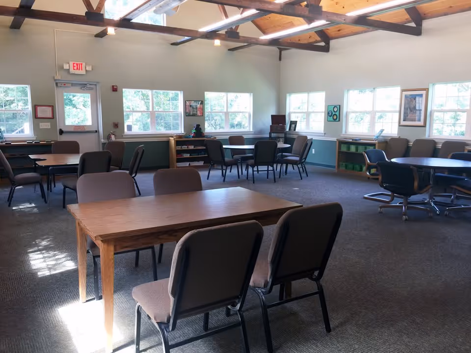 Large bright activity/dining room with multiple tables and chairs, windows along the walls, and exposed wooden ceiling beams.