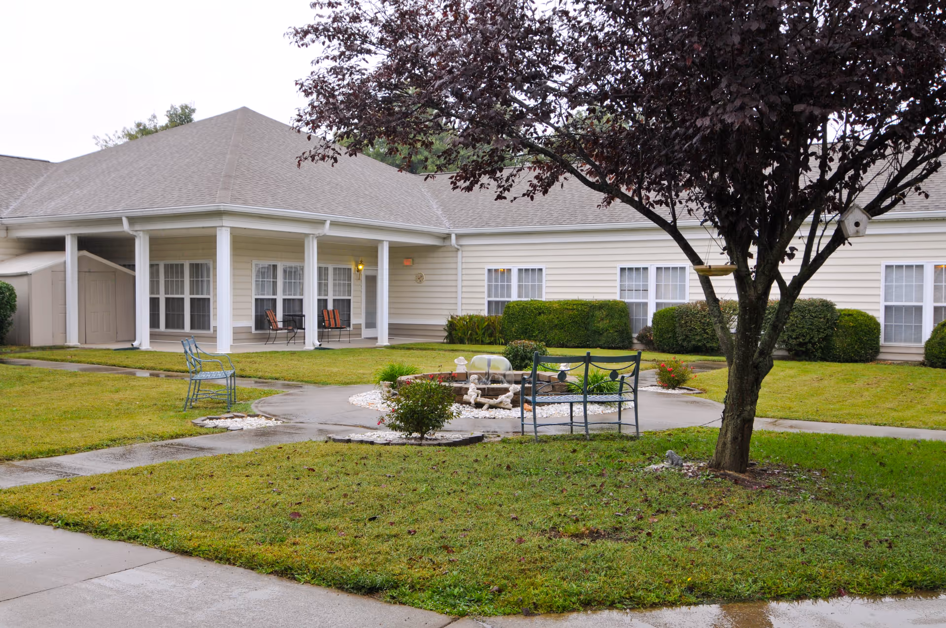 Outdoor courtyard area of a senior living facility with a tree, benches, a small fountain, and a building with multiple windows and a covered porch in the background.