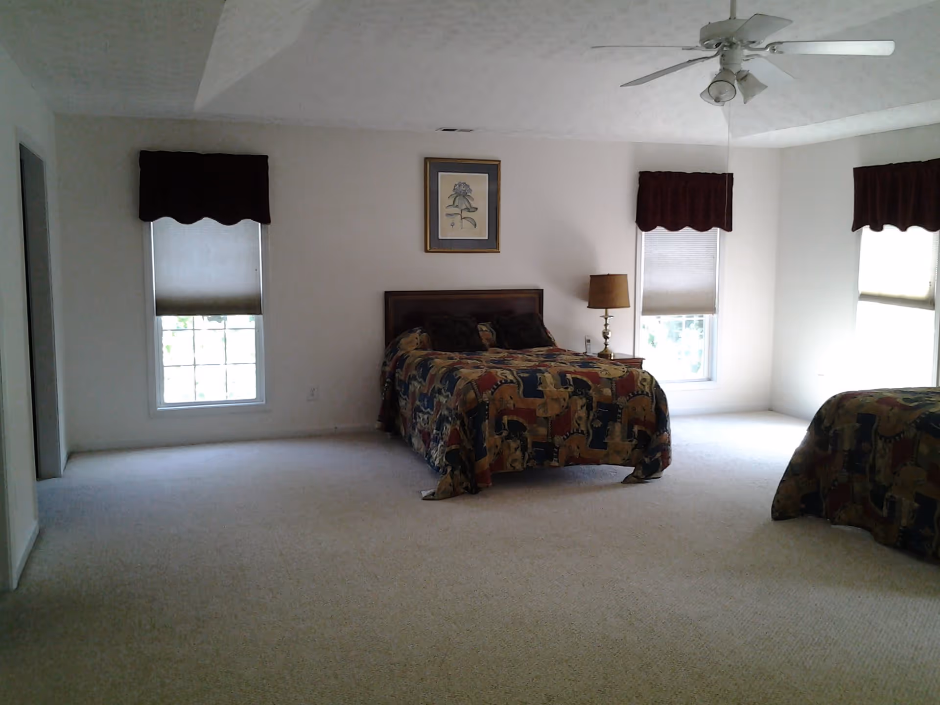 A spacious bedroom with two beds covered in patterned bedspreads, a nightstand with a lamp and phone, three windows with blinds and valances, a framed floral picture on the wall, and a ceiling fan with lights.