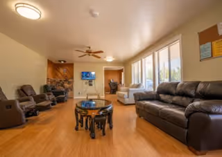 A spacious living room with wooden flooring, featuring a black leather sofa, several recliner chairs, a round wooden coffee table, a ceiling fan, a wall-mounted TV, and large windows allowing natural light to fill the room.