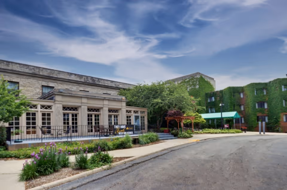Exterior view of a senior living facility named PA Peterson at the Citadel, showing a stone building with large windows, a patio area with chairs, lush greenery including ivy-covered walls, trees, and a clear blue sky with some clouds.