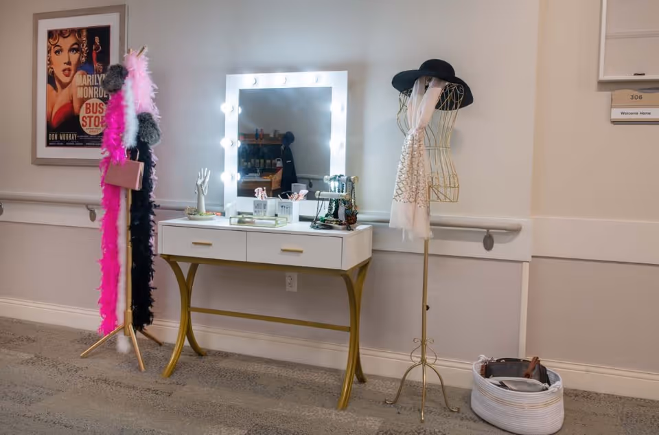 A vanity table with a lighted mirror, makeup and jewelry organizers, a mannequin stand with a black hat and white scarf, and a coat rack with colorful feather boas and a small purse. A framed Marilyn Monroe poster is on the wall to the left.