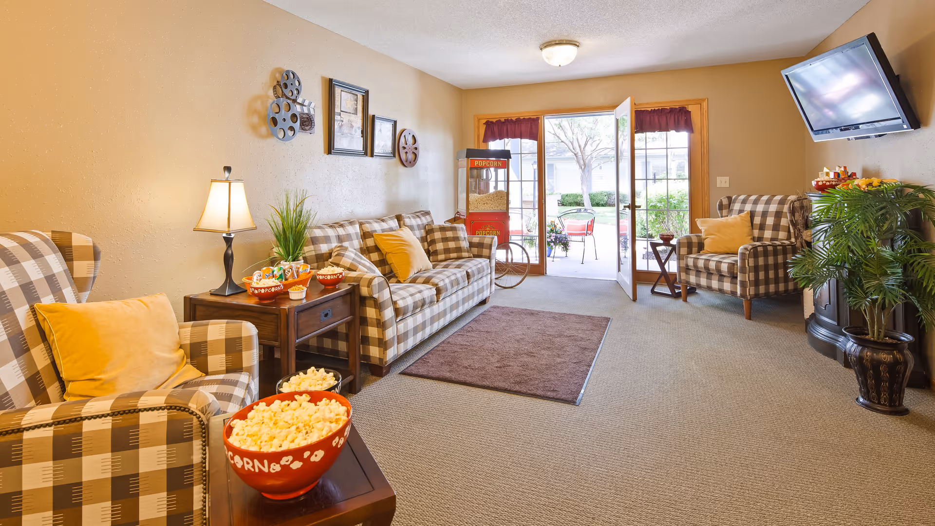 A cozy living room with plaid armchairs and a matching sofa, yellow accent pillows, a side table with a lamp and popcorn bowls, a popcorn machine near a glass door leading outside, a wall-mounted TV, and decorative plants.