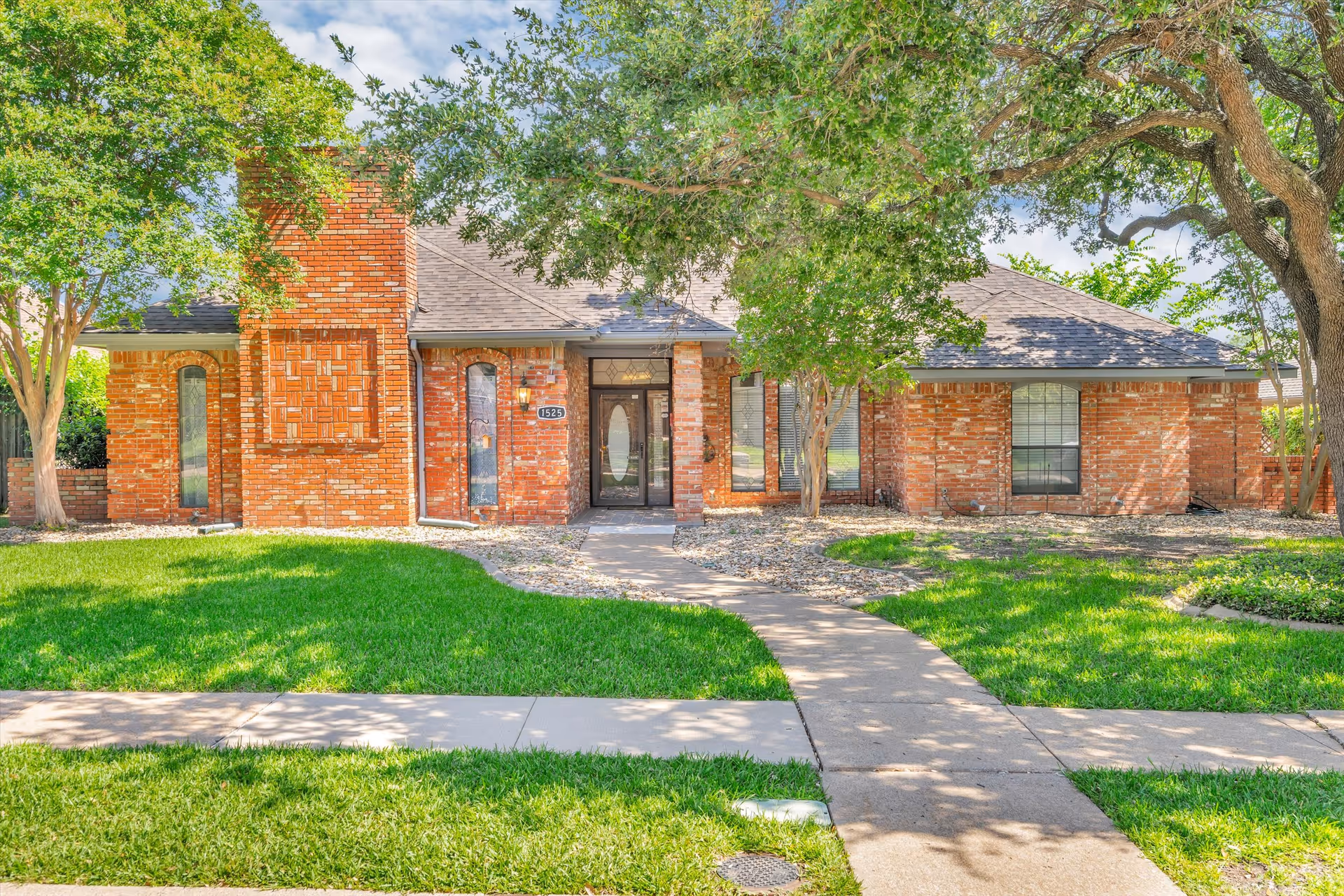 Front view of a single-story red brick house with a walkway, trees, and a green lawn.