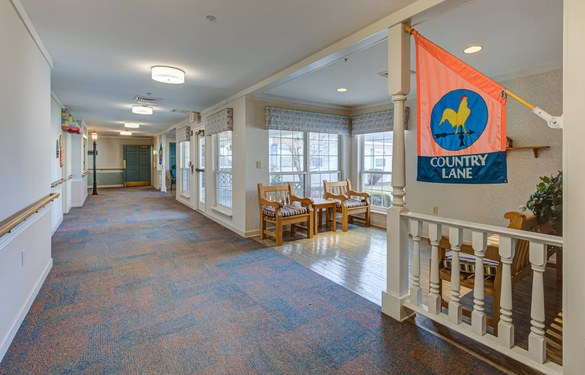 A long, carpeted hallway in a senior living facility with handrails on the walls and several ceiling lights. On the right side, there is a small seating area with two wooden chairs and a table near large windows. A colorful flag with a rooster and the words 'Country Lane' hangs above the seating area. The hallway leads to a green door at the far end.