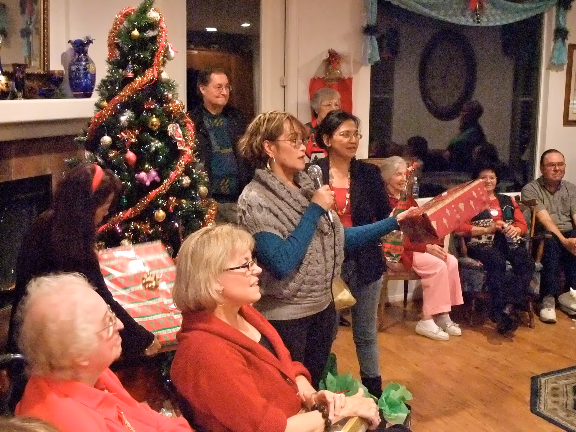 A group of people gathered in a living room decorated for Christmas with a decorated Christmas tree. One woman is holding a microphone and a wrapped gift, while others are seated or standing around, engaging in a holiday celebration.