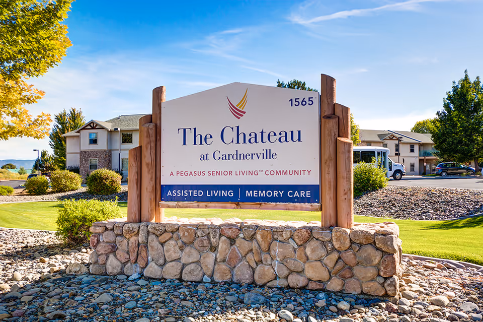 Outdoor view of the entrance sign for The Chateau at Gardnerville, a Pegasus Senior Living community offering assisted living and memory care. The sign is mounted on a stone base with wooden posts, surrounded by landscaped greenery and buildings in the background under a clear blue sky.