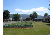 Well-kept lawn and garden beds with single-story cottage buildings overlooking a lake and distant hills under a blue sky.