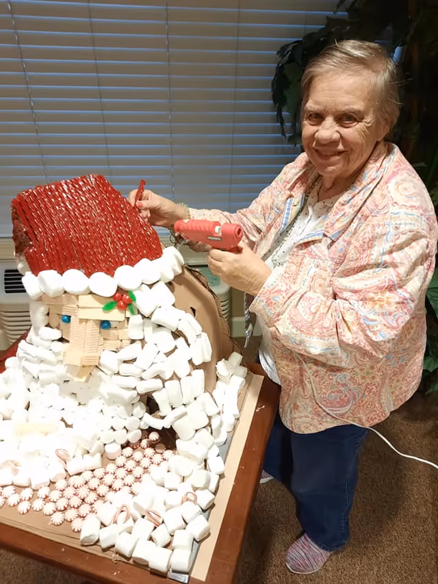 An elderly woman smiling while using a hot glue gun to assemble a large Santa Claus decoration made of various craft materials including marshmallows, peppermint candies, and red rope. She is standing indoors next to a table with the decoration on it, with window blinds and a plant in the background.
