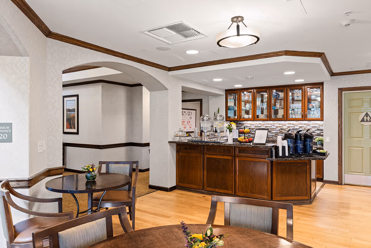 Interior view of a senior living facility dining area with wooden tables and chairs, a refreshment counter with water dispensers, coffee pots, cups, and a cabinet with glassware. The room has light-colored walls with a subtle pattern, wood trim, and a ceiling light fixture. A hallway and a door with a restroom sign are visible in the background.