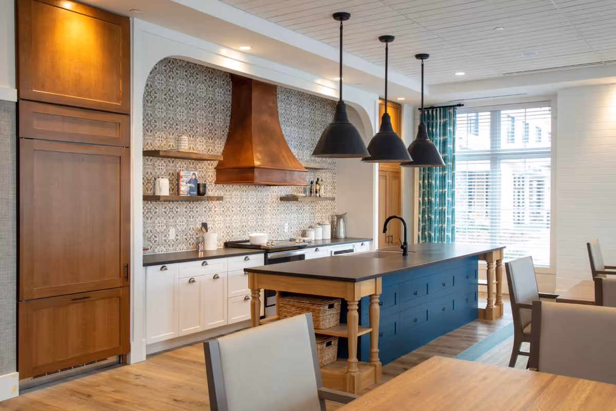 A modern kitchen with a large island featuring a dark countertop and blue cabinetry. The kitchen has white lower cabinets, wooden upper cabinets, and a copper range hood above the stove. Three black pendant lights hang above the island. There are shelves with decorative items on the tiled backsplash wall. The room has large windows with white blinds and teal curtains, and wooden dining tables and chairs are partially visible.