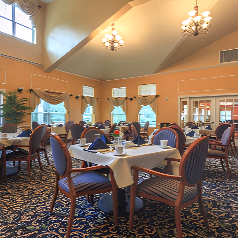Spacious dining room with round tables set with white linens and place settings, upholstered chairs, chandeliers, and large windows.