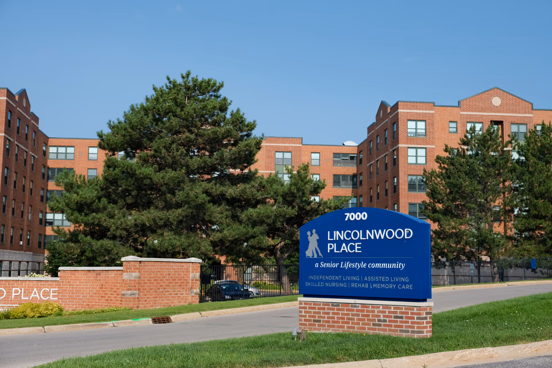 Exterior view of Lincolnwood Place, a senior lifestyle community, showing a large brick building with multiple windows behind a blue sign with white text listing services like independent living, assisted living, skilled nursing, rehab, and memory care. The scene includes green trees, grass, and a clear blue sky.