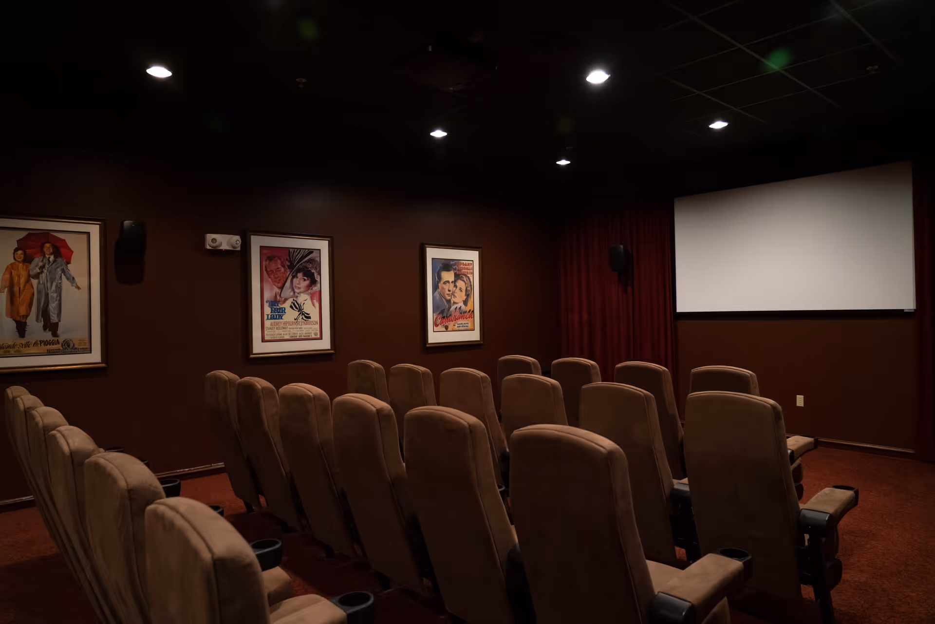 A small movie theater room with three rows of brown cushioned seats facing a large white projection screen. The walls are dark brown and decorated with framed vintage movie posters. The ceiling has recessed lighting.