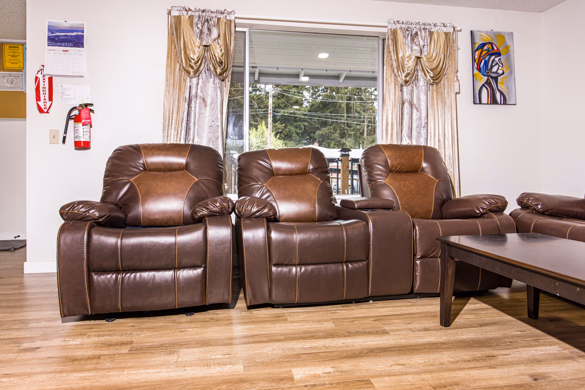 A living room with three brown leather recliner chairs arranged side by side in front of a large window with beige and gold curtains. There is a wooden coffee table in front of the chairs. On the wall to the left, there is a fire extinguisher and a calendar. On the right wall, there is a colorful painting of a person wearing a headwrap.