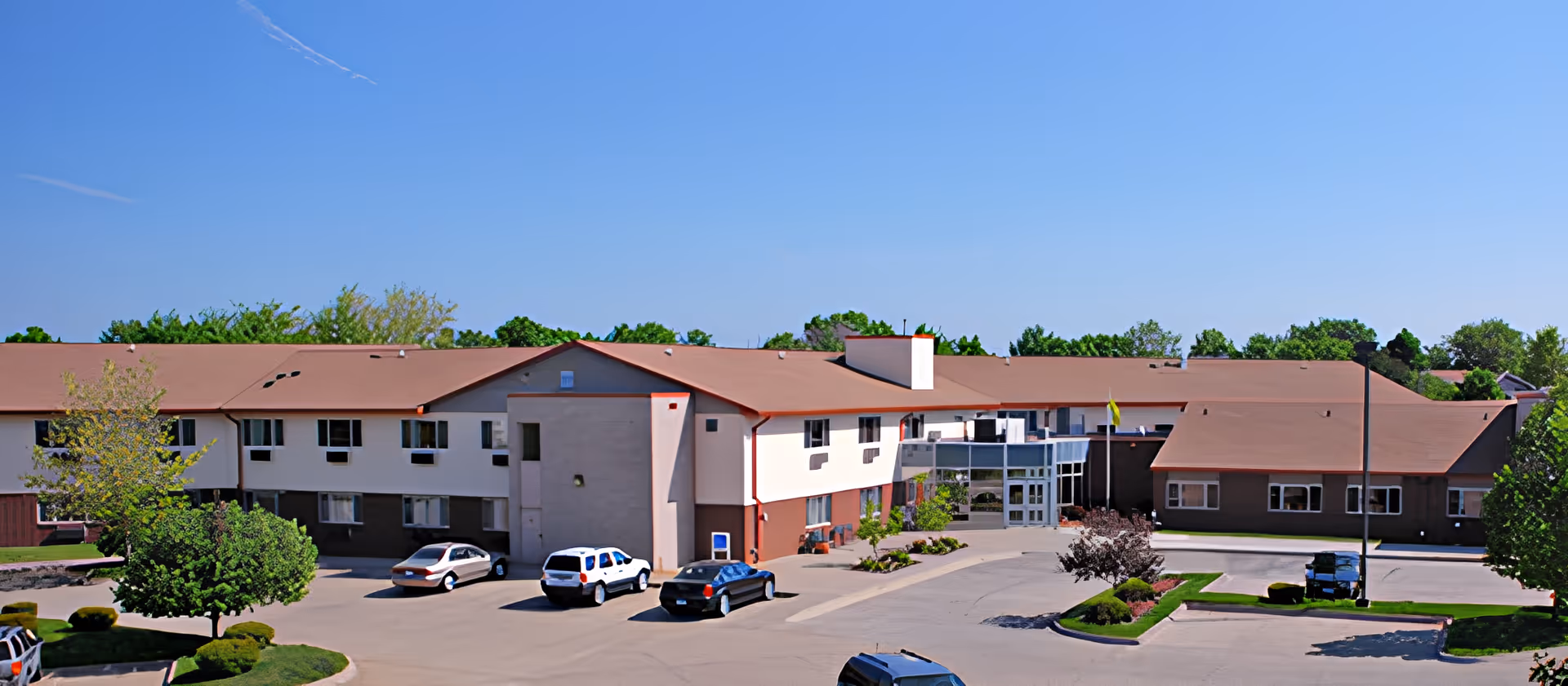 Front exterior of a two-story senior living facility with a parking lot, several cars, and landscaped grounds under a clear blue sky.