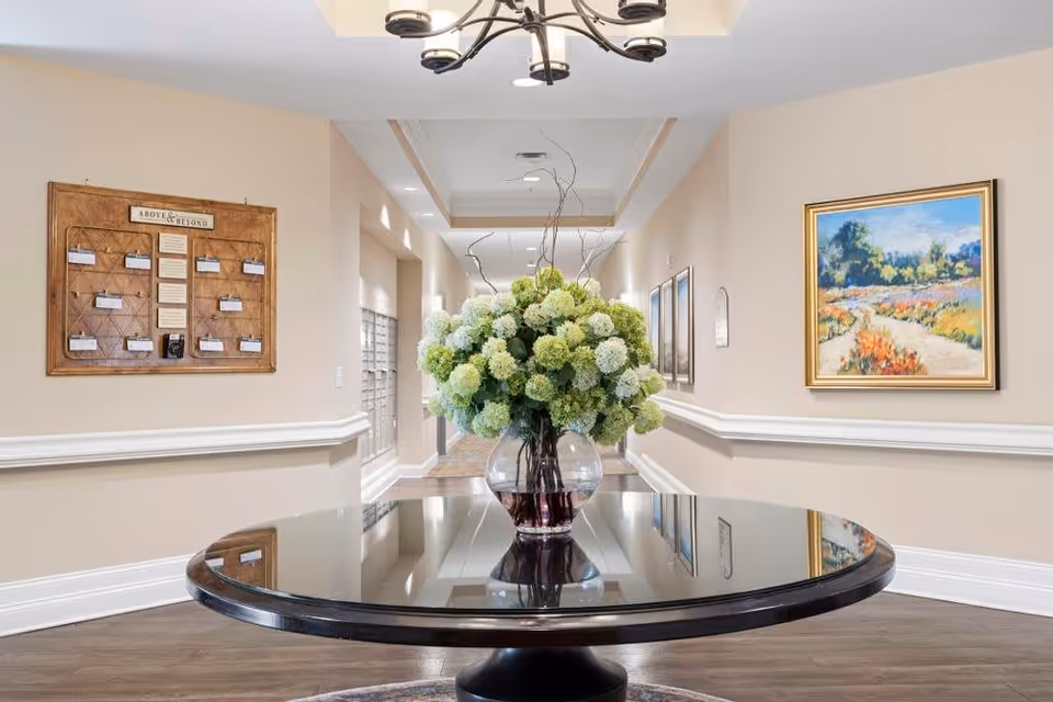 A hallway in a senior living facility with a round black table in the center holding a large glass vase filled with green and white hydrangea flowers. The walls are beige with white trim and decorated with framed artwork and a bulletin board. The floor is wooden, and there is a modern chandelier hanging from the ceiling.