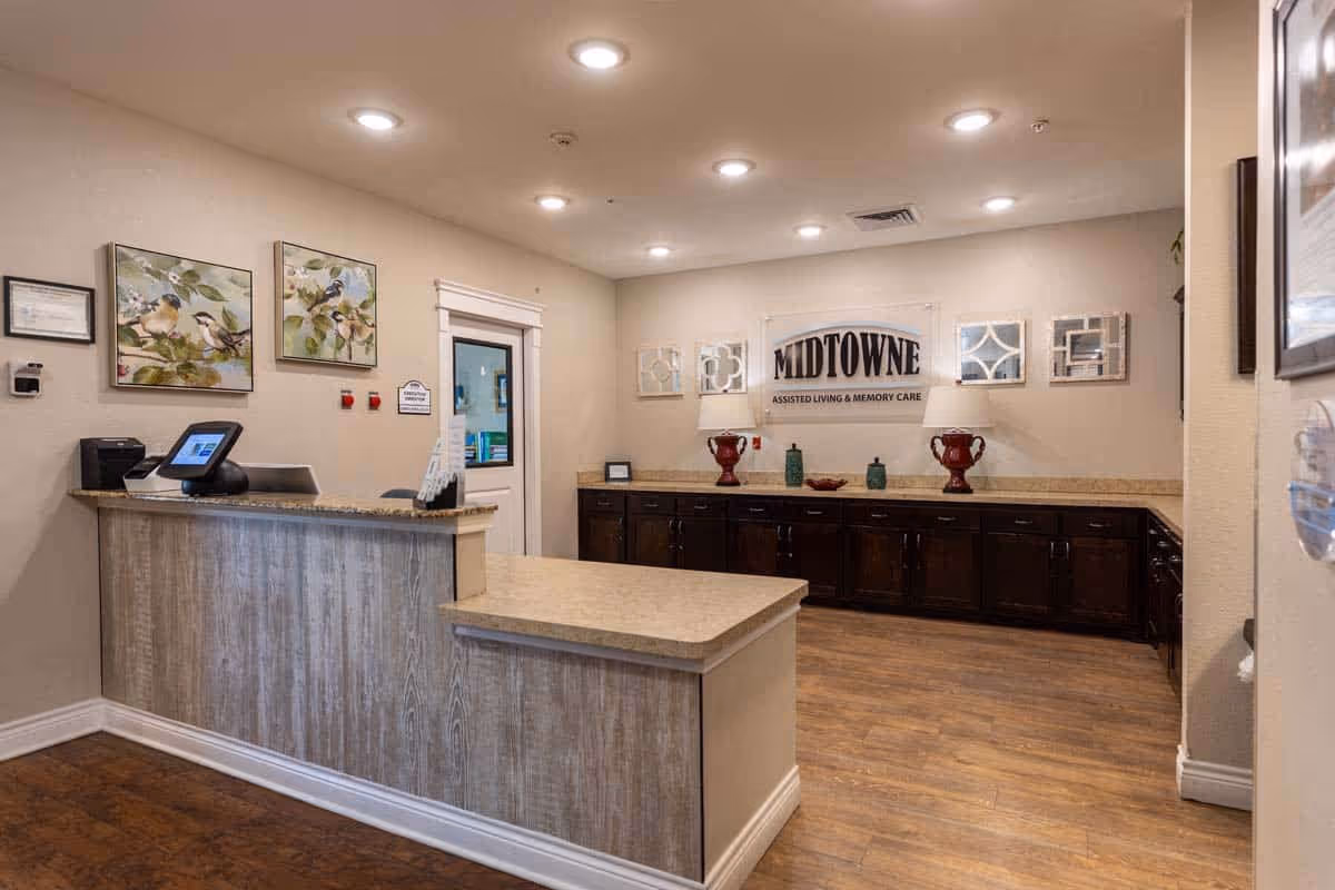 Reception area of Midtowne Assisted Living and Memory Care with a wooden front desk, a computer, decorative bird paintings on the wall, and a sign displaying the facility's name above a counter with decorative items.