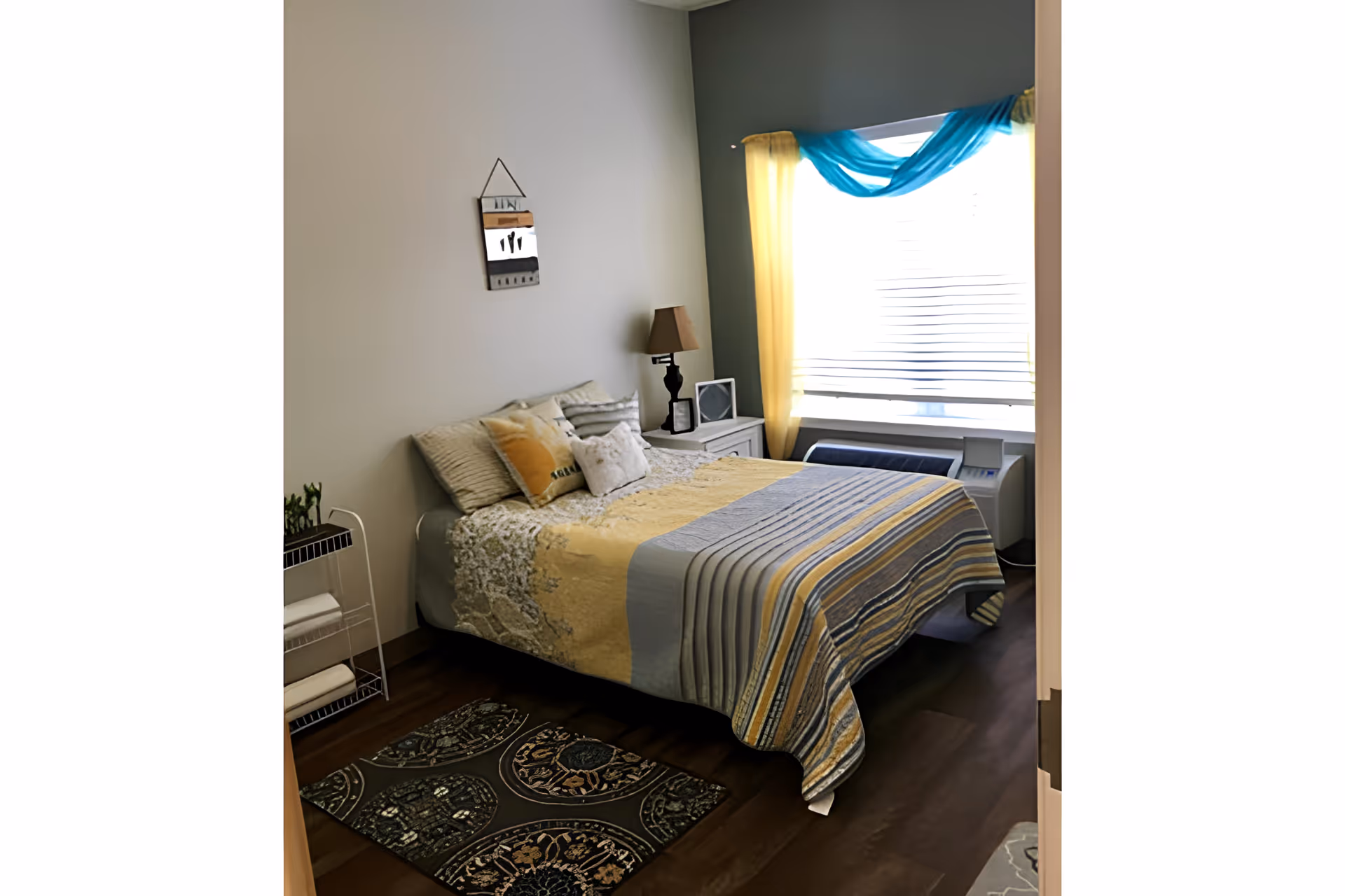 A cozy bedroom with a bed covered in a yellow, gray, and blue striped quilt. The bed has multiple pillows and is positioned against a light gray wall. There is a small white nightstand with a lamp and a framed picture next to the bed. A window with yellow curtains and a blue sheer drape allows natural light into the room. A decorative rug is placed on the wooden floor near the bed.