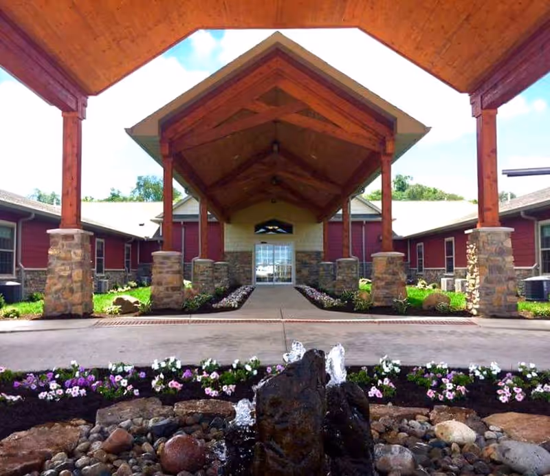 Covered porte-cochere with wooden beams and stone pillars leading to the building entrance, with a small fountain and flower beds in the foreground.