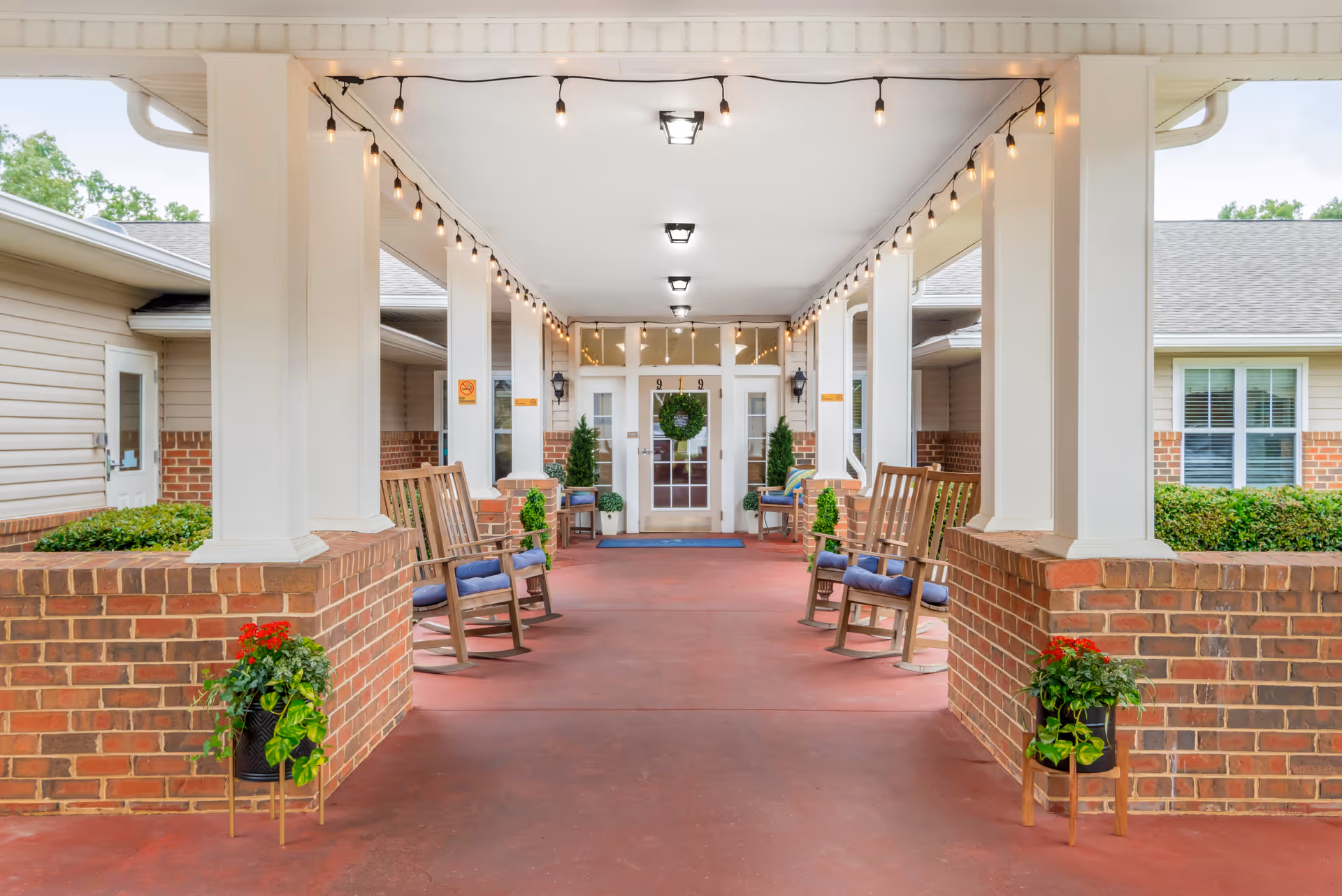 Covered entrance area of a senior living facility with brick pillars and walls, wooden rocking chairs with blue cushions arranged on both sides, string lights hanging from the ceiling, potted plants, and a glass door decorated with a green wreath at the far end.