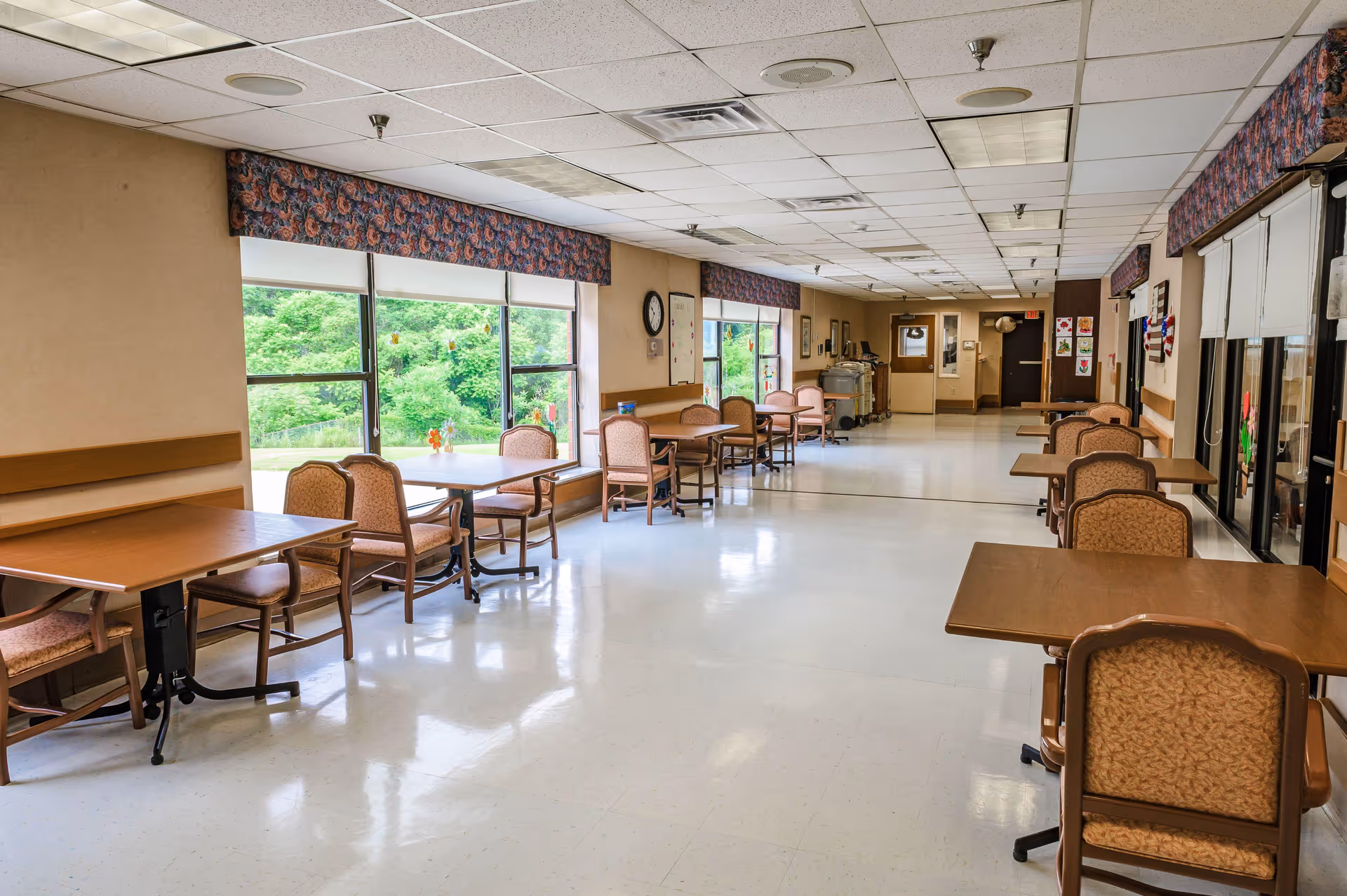 A bright dining area in a senior living facility with multiple wooden tables and cushioned chairs arranged along large windows that show green trees outside. The room has a tiled ceiling with fluorescent lights and floral valances above the windows. The floor is light-colored and shiny, and the walls are beige with some decorations and a clock visible in the background.