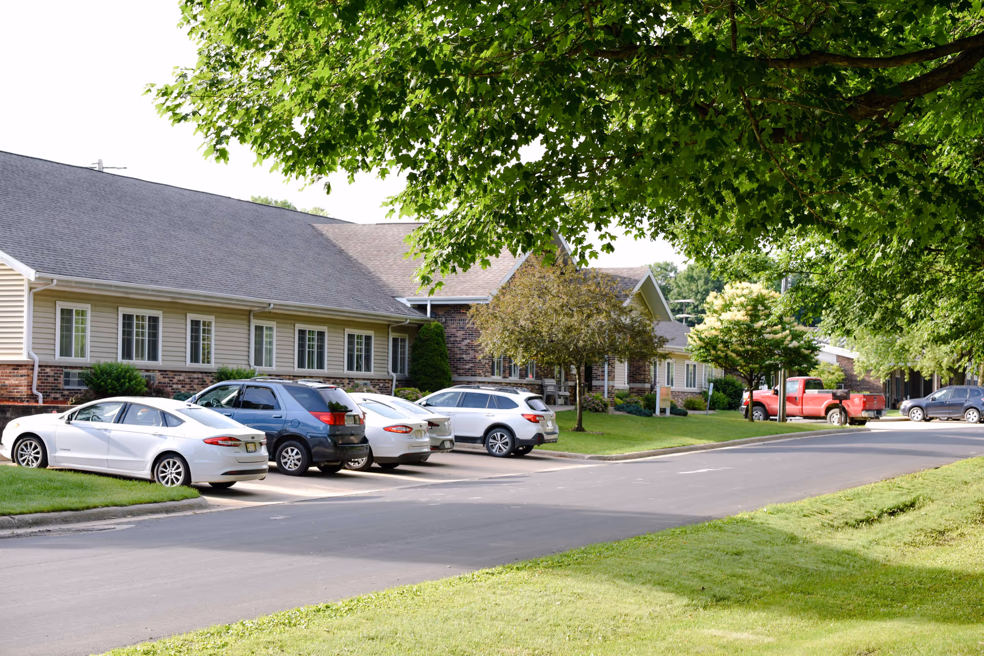 Exterior view of Maple Crest Campus showing a single-story building with beige siding and brick accents. Several cars are parked in front along a paved driveway, with green grass and trees providing shade and a natural setting.