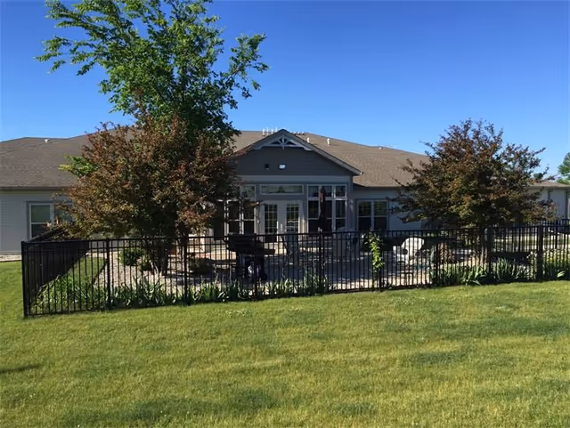 View of a single-story building with a fenced patio area in front, featuring trees, outdoor furniture, and a well-maintained lawn under a clear blue sky.