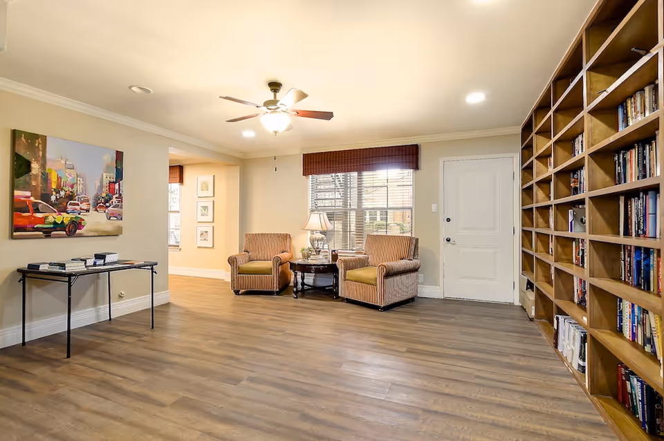 Bright common room with two armchairs and a side table, a large built-in bookshelf, artwork on the wall, and wood flooring.