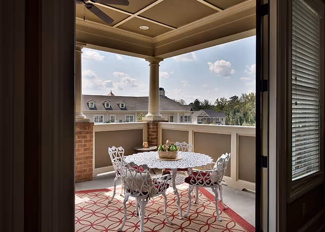 View of a covered outdoor balcony with a white ornate metal table and four matching chairs with red cushions, a patterned outdoor rug underneath, and a basket centerpiece on the table. The balcony overlooks a building with dormer windows and a partly cloudy sky.