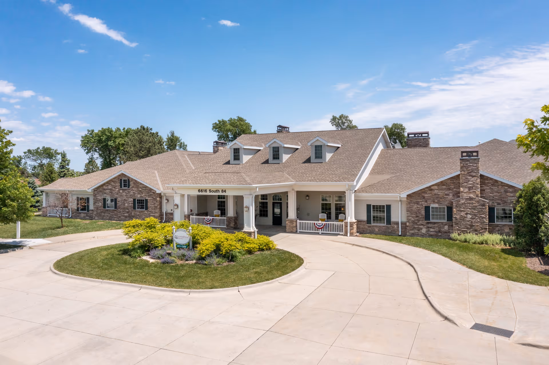 Front exterior view of a single-story senior living facility building with a circular driveway, manicured lawn, and shrubs under a blue sky with scattered clouds.