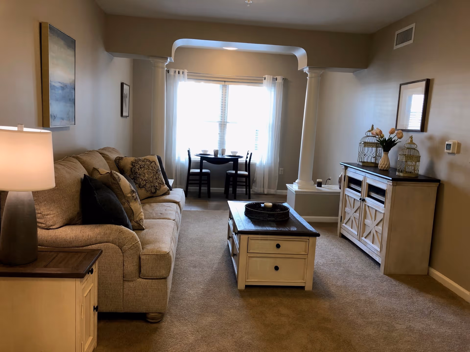 A cozy living room with a sofa, coffee table, sideboard, and a small dining table by a window framed by decorative columns.