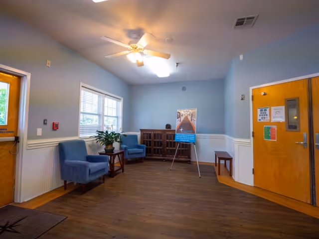 A small waiting area with two blue armchairs, a wooden side table with a potted plant, a wooden cabinet, and a standing sign. The room has light blue walls with white wainscoting, a ceiling fan with lights, and wooden doors on either side.