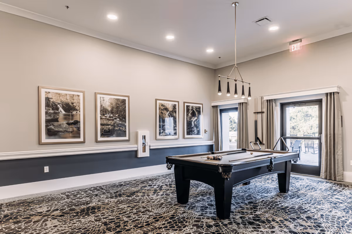 A well-lit game room with a pool table in the center, featuring a modern hanging light fixture above it. The room has a patterned carpet, four framed nature photographs on the wall, and two glass doors with curtains leading outside. A pool cue rack is mounted on the wall near the doors.