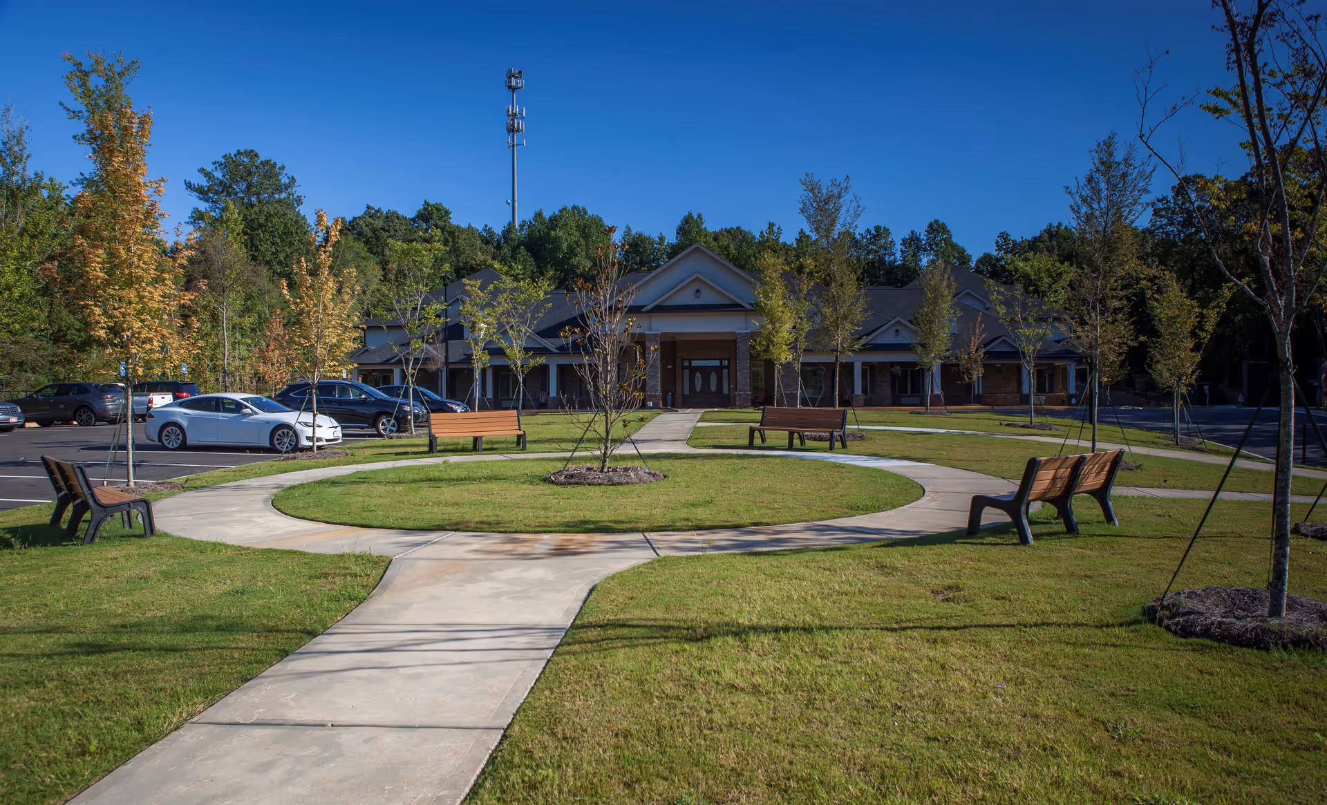 Outdoor view of a senior living facility with a circular concrete pathway surrounded by green grass and benches. Several young trees are planted around the pathway, and a parking lot with cars is visible to the left. The building is a single-story structure with a peaked roof and a covered entrance, set against a backdrop of tall trees under a clear blue sky.