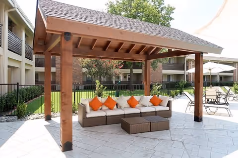 Outdoor seating area at MontClair Estates featuring a wooden pergola with a cushioned sectional sofa adorned with orange and beige pillows, set on a tiled patio with additional tables and chairs nearby, surrounded by greenery and apartment buildings.