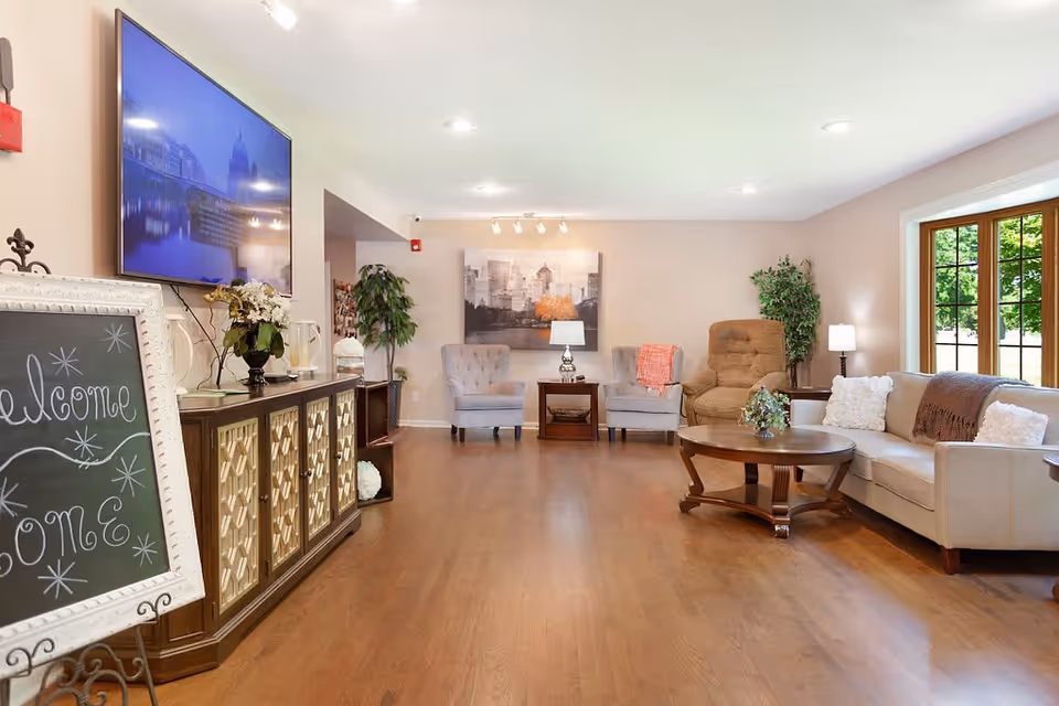 A bright and welcoming living room area in a memory care home with wooden flooring, a beige sofa with white and brown pillows, a round wooden coffee table with a small plant, two gray armchairs, a brown recliner, a side table with a lamp, a large window showing greenery outside, a large wall-mounted TV, a decorative cabinet with flowers, and a chalkboard sign that says 'Welcome Home'.