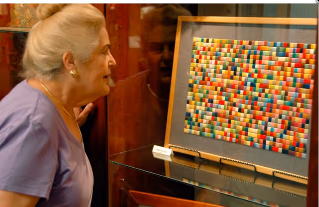 An elderly woman with gray hair tied back is closely observing a colorful, rectangular mosaic artwork displayed in a glass case at Asbury Methodist Village.