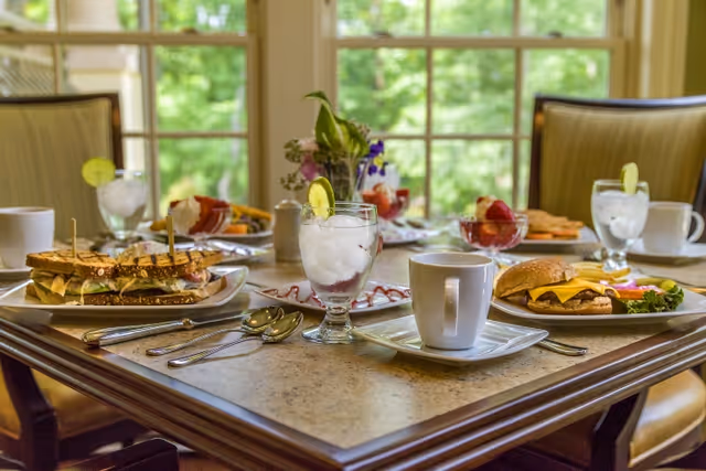 A dining table set with plates of sandwiches, cheeseburgers, glasses of water with lemon slices, coffee cups, and bowls of fruit salad, with a window showing green trees in the background.